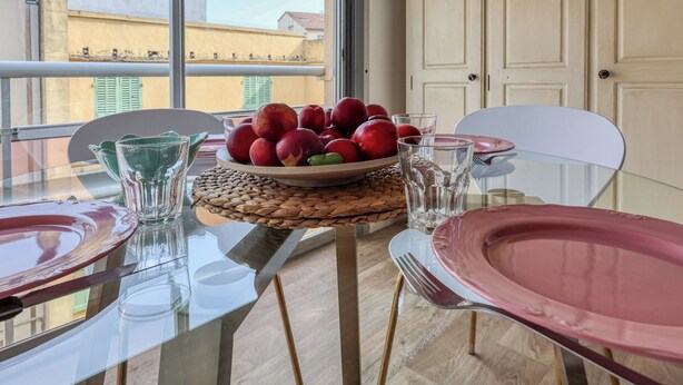 Een eettafel met een schaal fruit, glazen en borden, op een balkon met uitzicht op gebouwen.