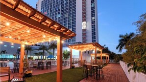 A hotel with a covered outdoor area featuring tables and chairs, a wooden deck, and a tall building in the background. - 24th Floor City View. Luxury Downtown Apartment, Pool, Spa, Gym, Free Parking. (Fort Myers)