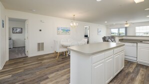 A modern kitchen with white cabinetry, a central island, and a dining area with white chairs. - Linens Supplied Rehoboth Beach Condo Walk to Shops and Restaurants (Rehoboth Beach)