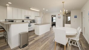 A modern kitchen with white cabinetry, a central island, and a dining area with white chairs. - Linens Supplied Rehoboth Beach Condo Walk to Shops and Restaurants (Rehoboth Beach)