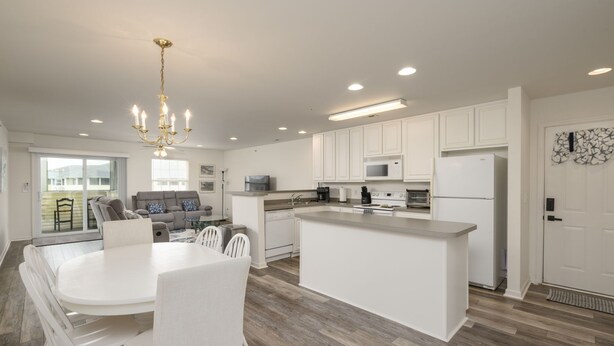A modern kitchen with white cabinetry, a central island, and a dining area with white chairs. - Linens Supplied Rehoboth Beach Condo Walk to Shops and Restaurants (Rehoboth Beach)
