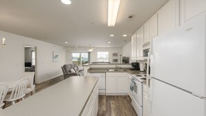 A modern kitchen with white cabinetry, a central island, and a dining area with white chairs. - Linens Supplied Rehoboth Beach Condo Walk to Shops and Restaurants (Rehoboth Beach)