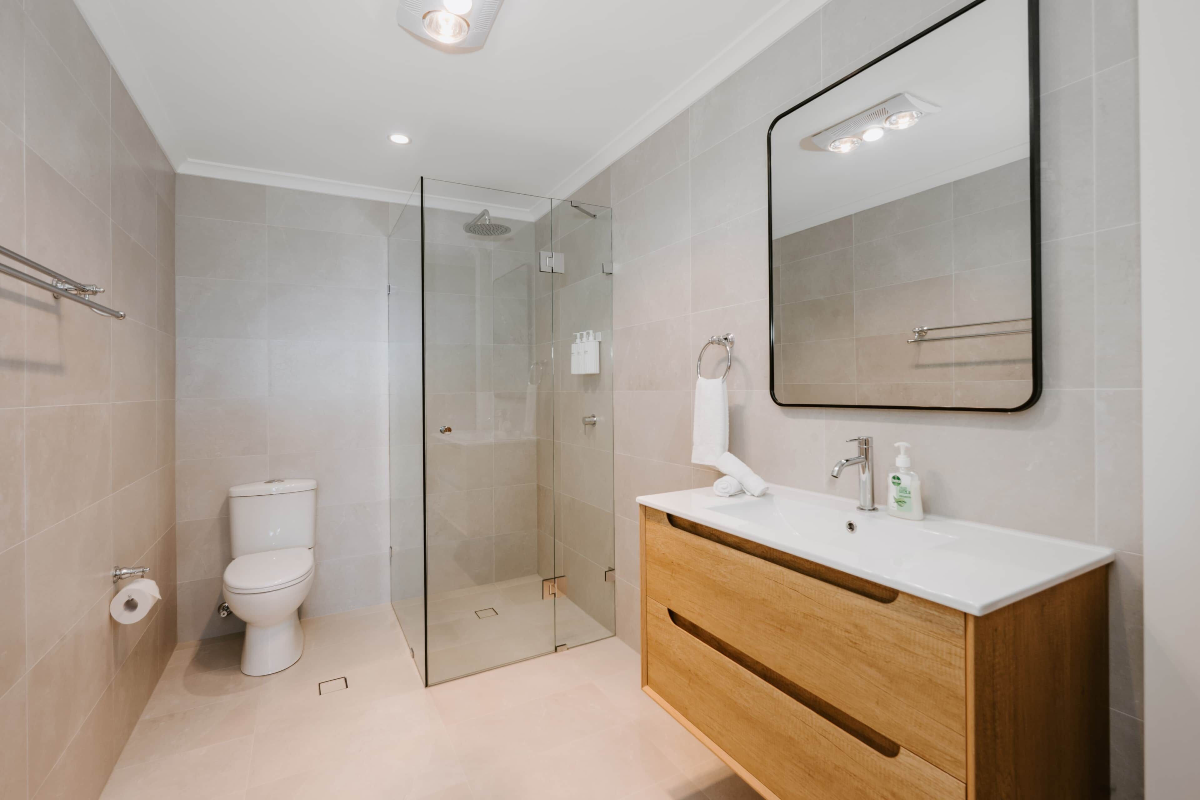 Een moderne badkamer met een douchecabine van glas, een wit toilet en een houten wastafelmeubel met spiegel.