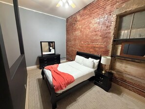 Une chambre avec un mur en briques, un lit recouvert d’une couette blanche et d’un plaid rouge, une commode, une table de chevet, un miroir et une étagère fixée au mur.