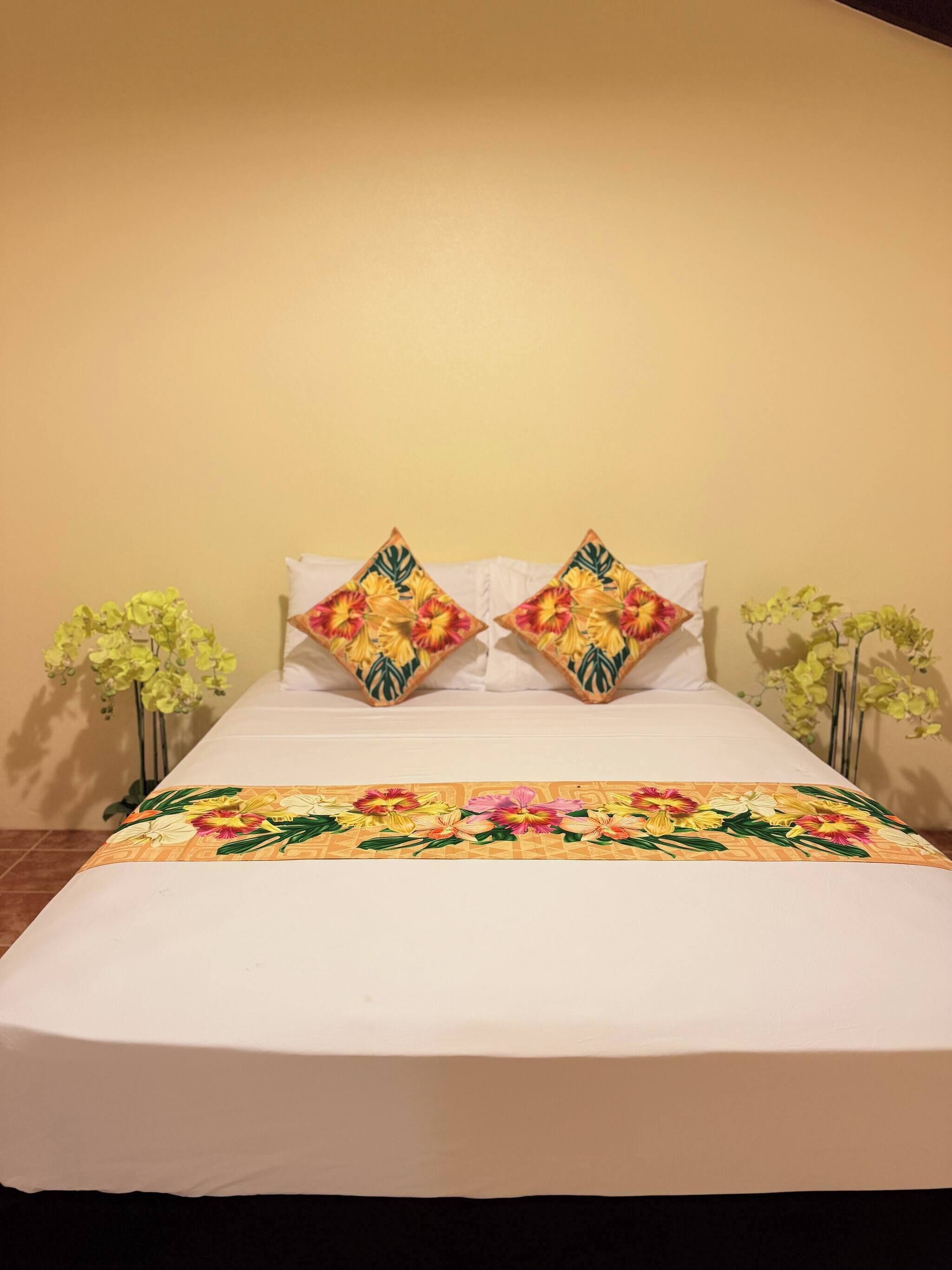 A bed with a floral bedspread, two pillows with a tropical pattern, and two potted plants on either side.