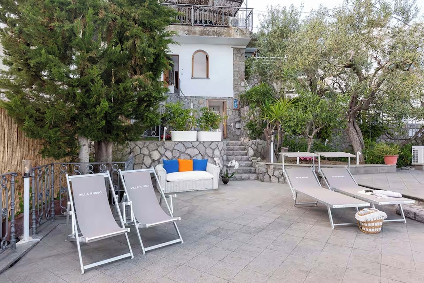 A villa with outdoor seating, a stone wall, and a staircase leading to a balcony.