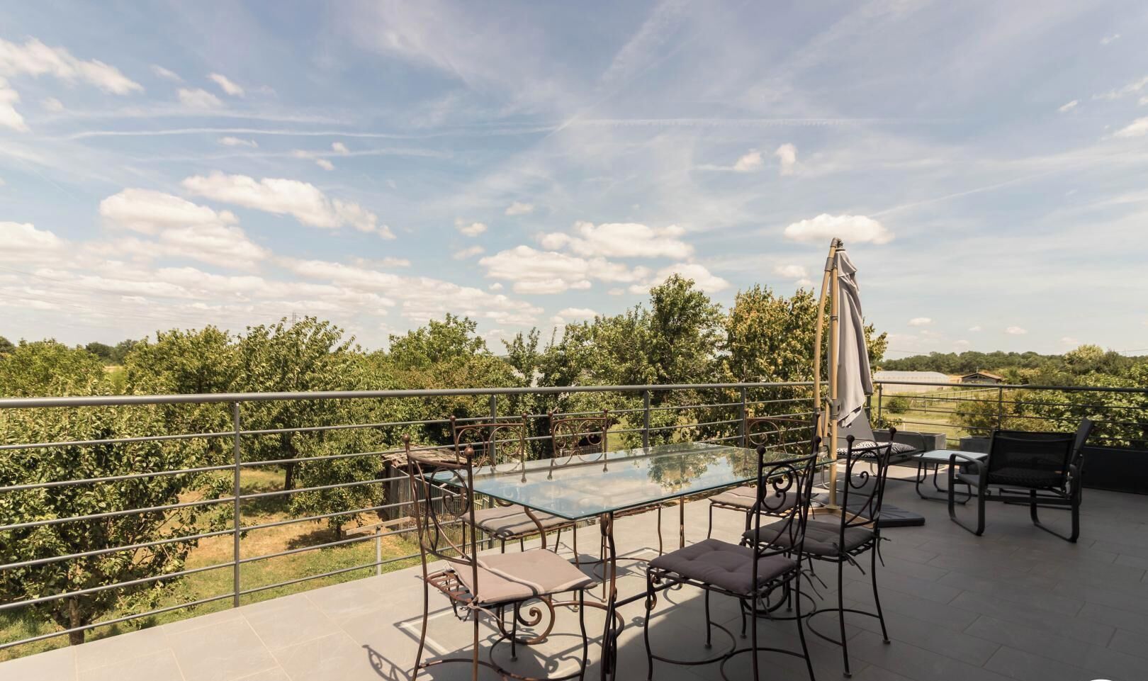 Een glazen tafel met metalen stoelen en een parasol op een balkon met een metalen hekwerk.