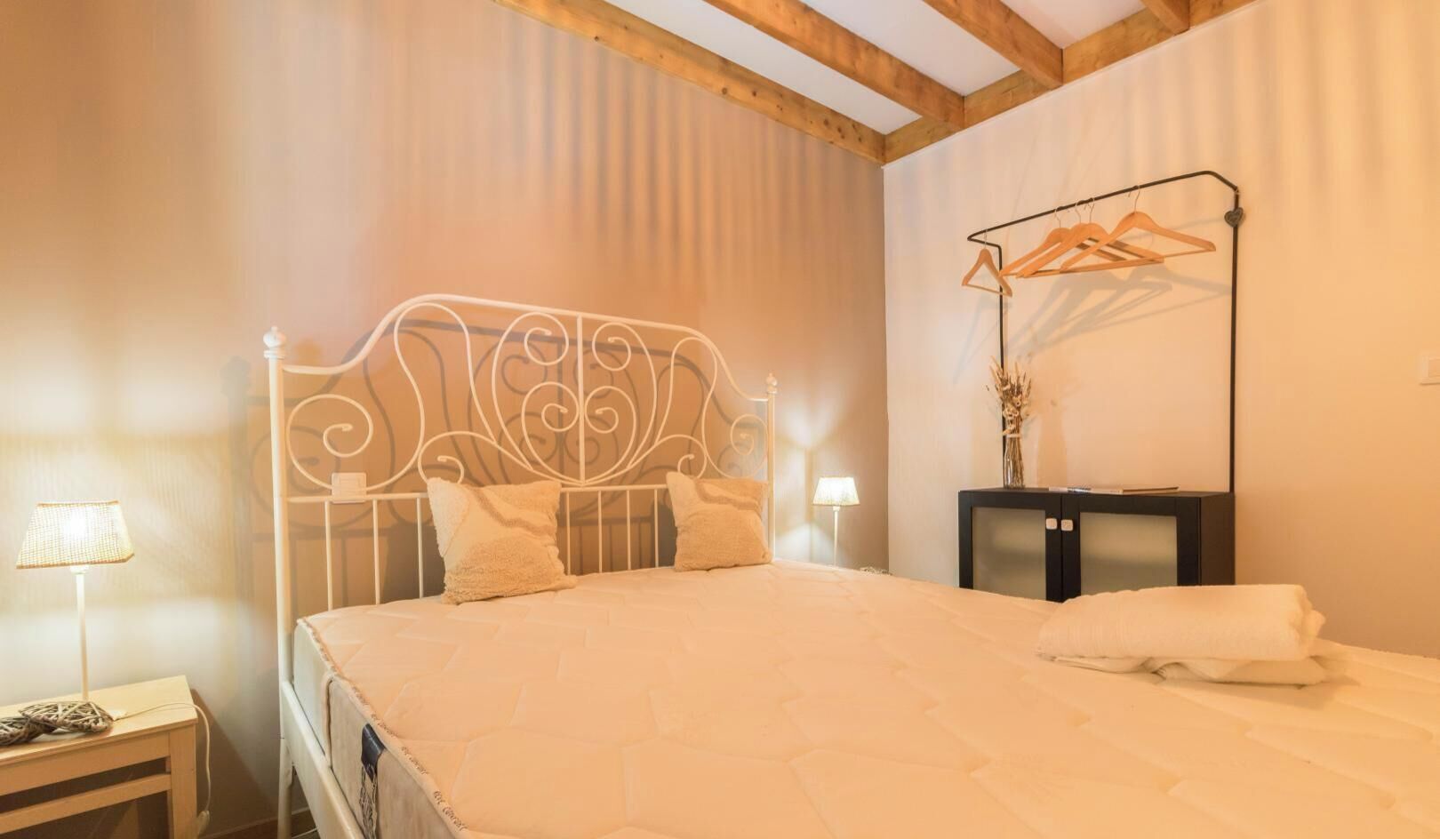 Un lit bien fait, avec une tête de lit en métal, une table de chevet avec une lampe et un portant à vêtements suspendu.