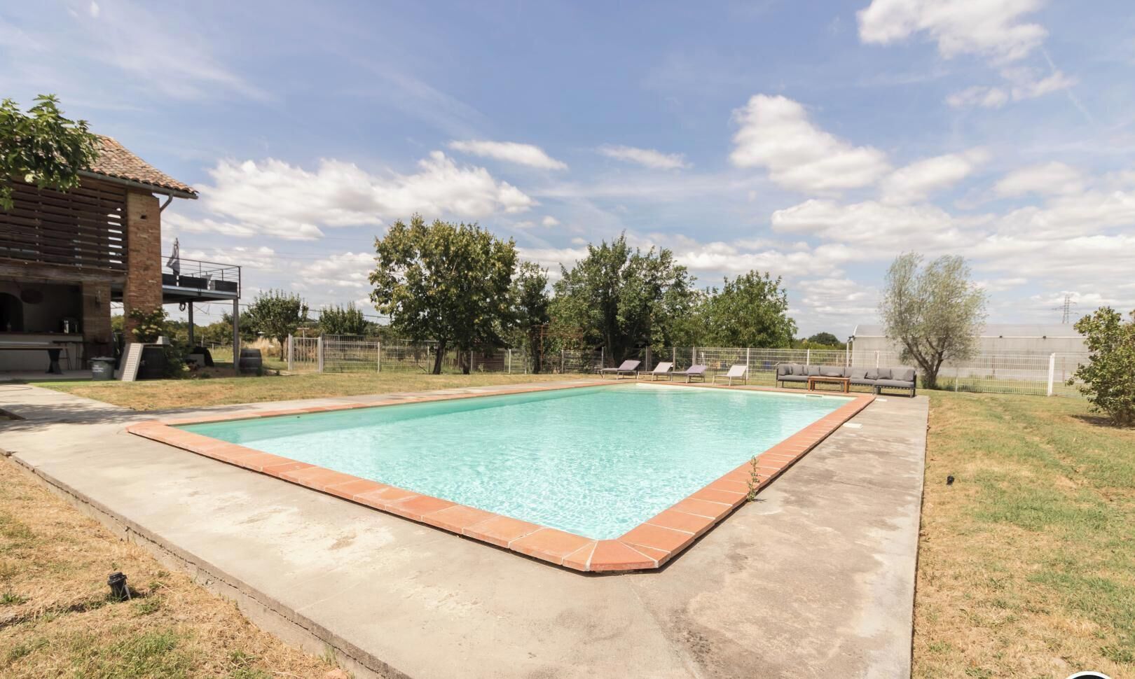 Une piscine rectangulaire dont les bords sont recouverts de carreaux, entourée d’une pelouse et d’arbres.