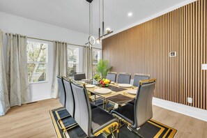 A dining area with a table set for six, surrounded by chairs with gold accents, a wooden slatted wall, and a window with curtains. - New! Ivy Luxury Home Near Airport And Downtown (East Point)