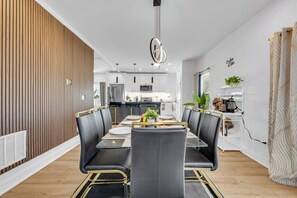 A modern dining area with a long table, black chairs with gold accents, and a central light fixture. - New! Ivy Luxury Home Near Airport And Downtown (East Point)