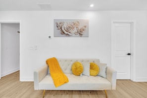 A white sofa with a yellow throw blanket and two yellow pillows, a white door, and a wall-mounted artwork of flowers. - New! Ivy Luxury Home Near Airport And Downtown (East Point)