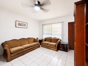 A living room with a brown sofa set, a ceiling fan, a window with blinds, and a wooden cabinet.