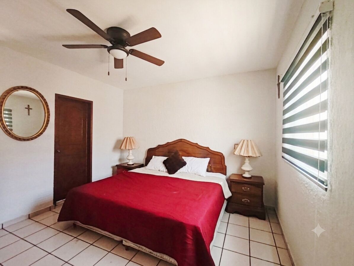 Une chambre à coucher comprenant un lit, deux tables de chevet, un ventilateur de plafond, une fenêtre avec des stores, une porte et un miroir mural.