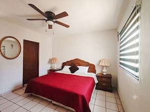 A bedroom with a bed, two bedside tables, a ceiling fan, a window with blinds, a door, and a mirror on the wall.