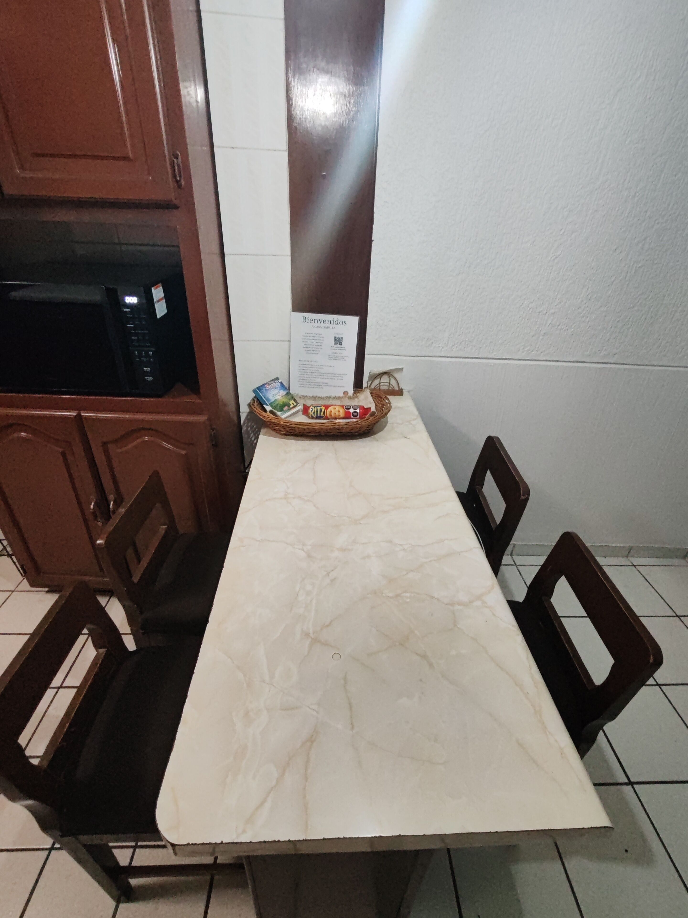 Une table à manger avec un plateau en marbre, entourée de quatre chaises, dans une pièce équipée d’un four à micro-ondes et agrémentée d’une pancarte de bienvenue.