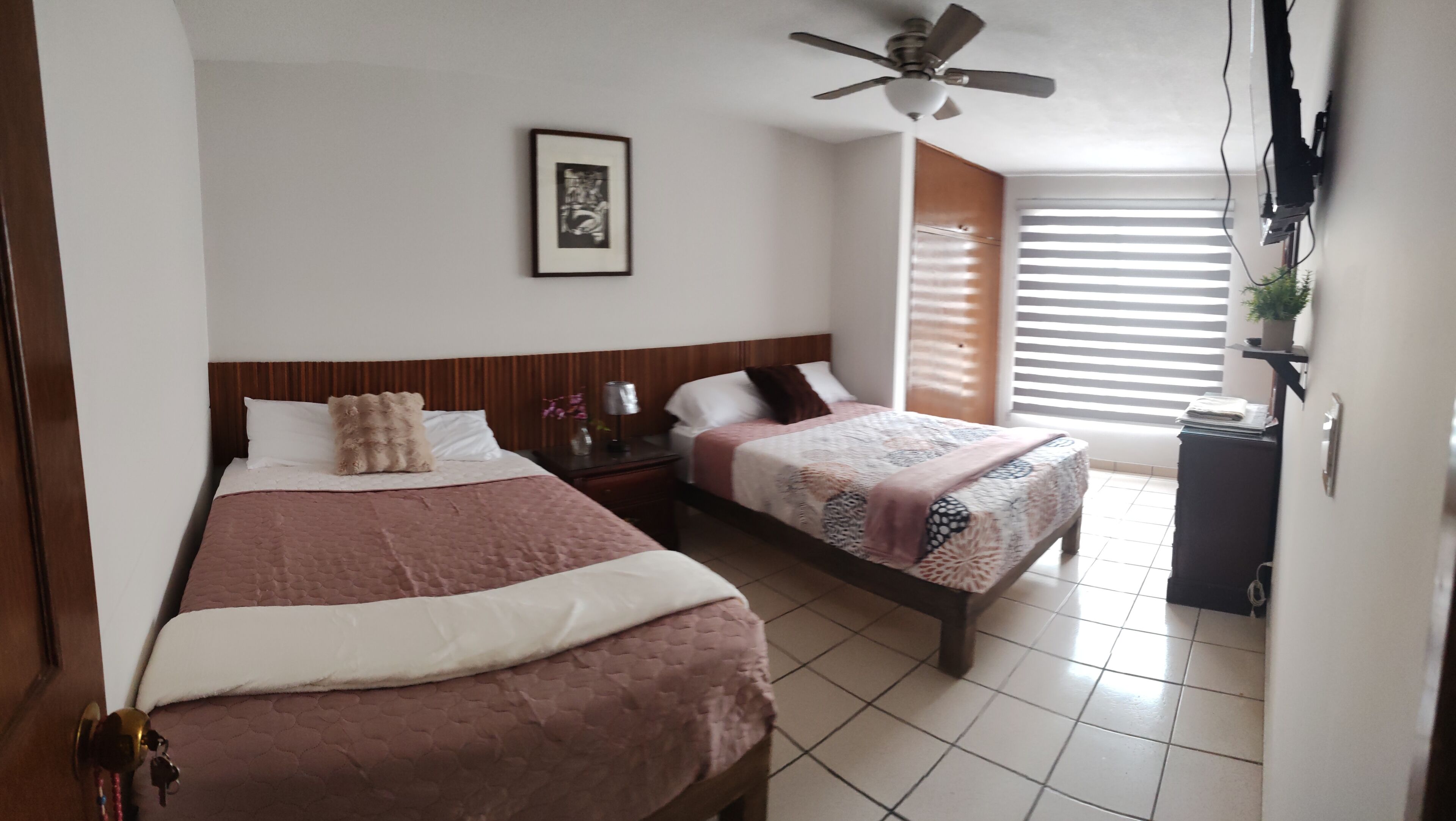 Een hotelkamer met twee bedden, een plafondventilator, een ingelijst beeld aan de muur en een raam met zonwering.