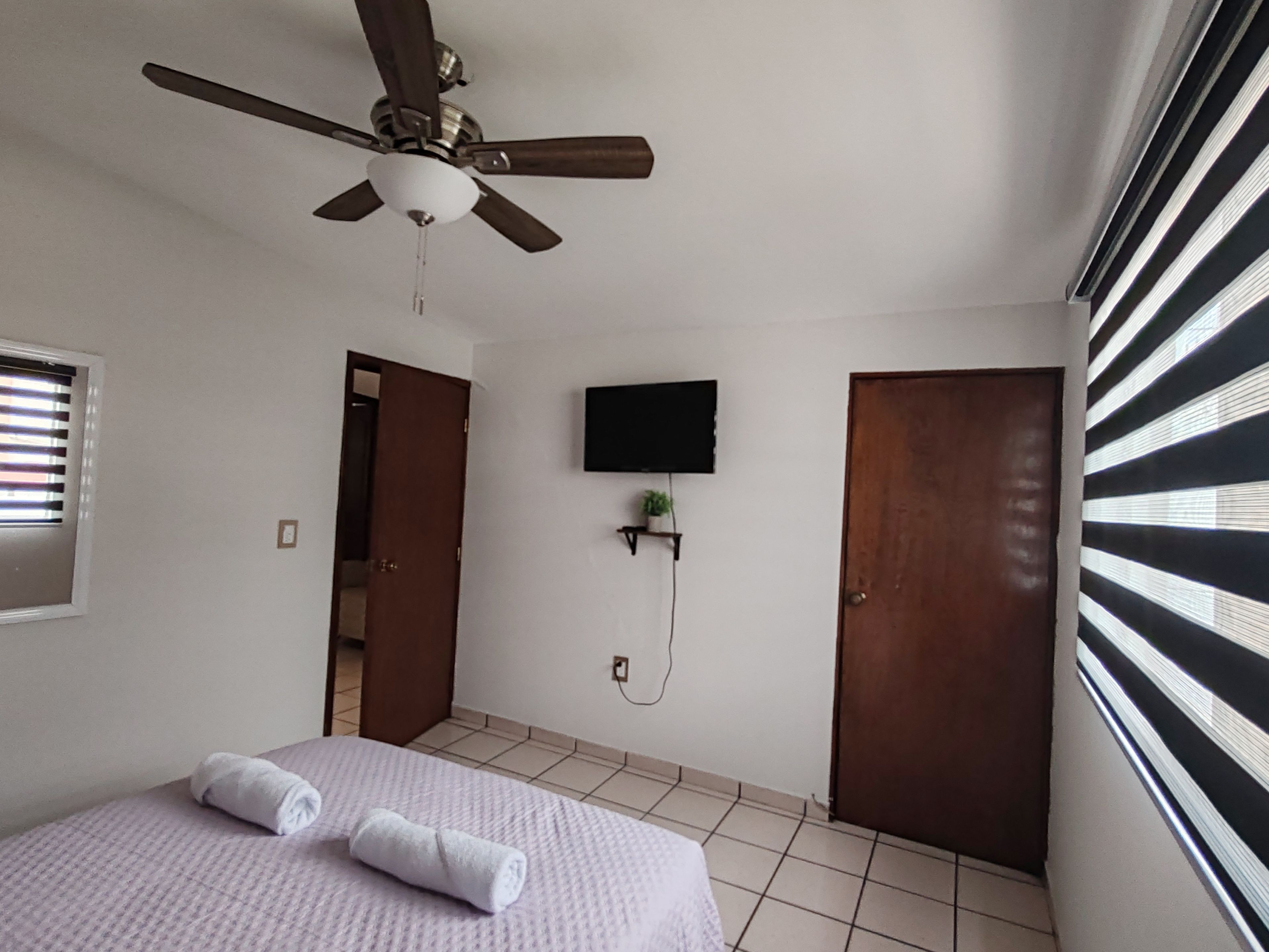 A hotel room with a bed, a ceiling fan, a TV mounted on the wall, and a door.