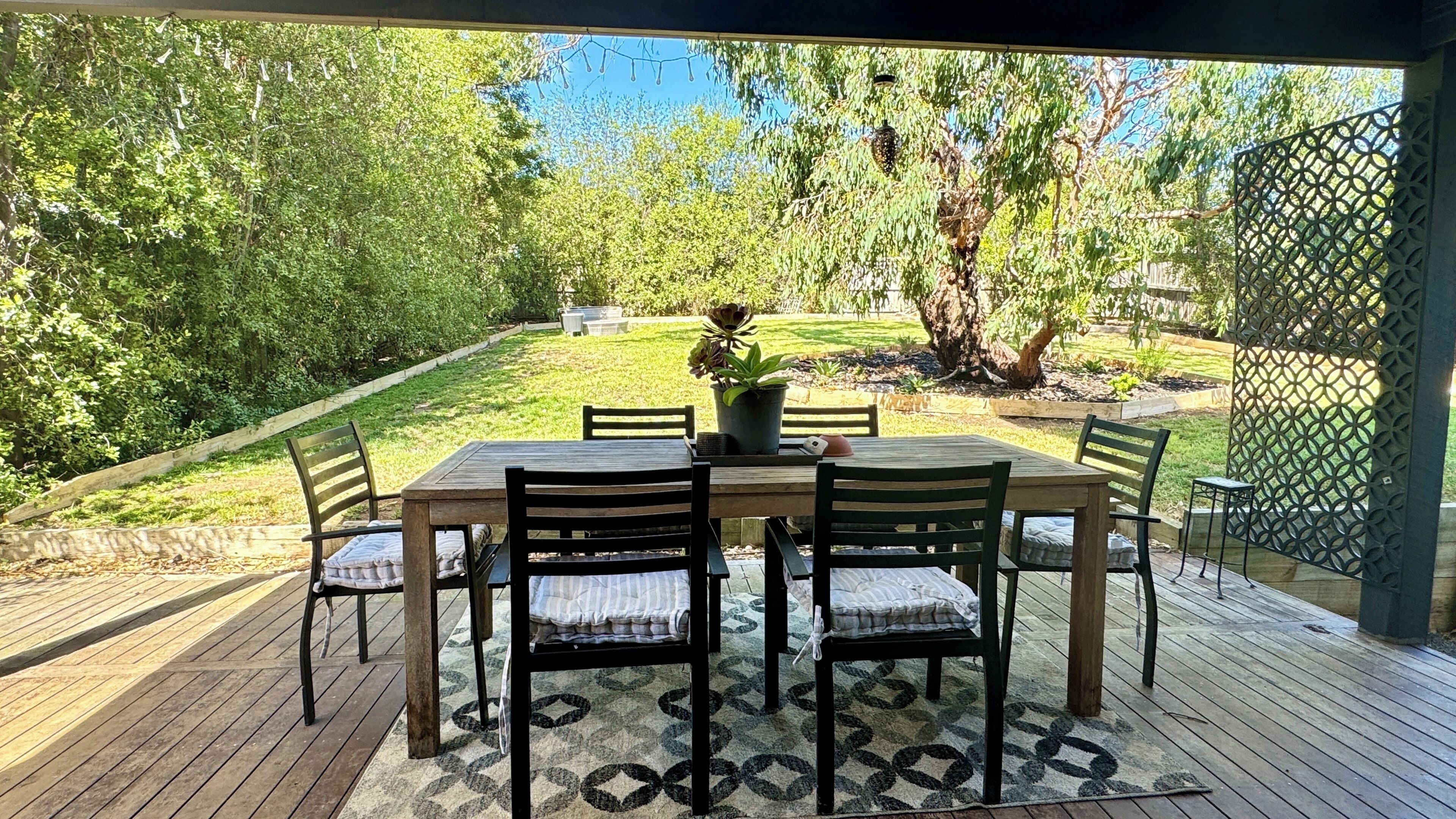 An outdoor patio with a wooden table and chairs, surrounded by a wooden deck and a view of a garden.