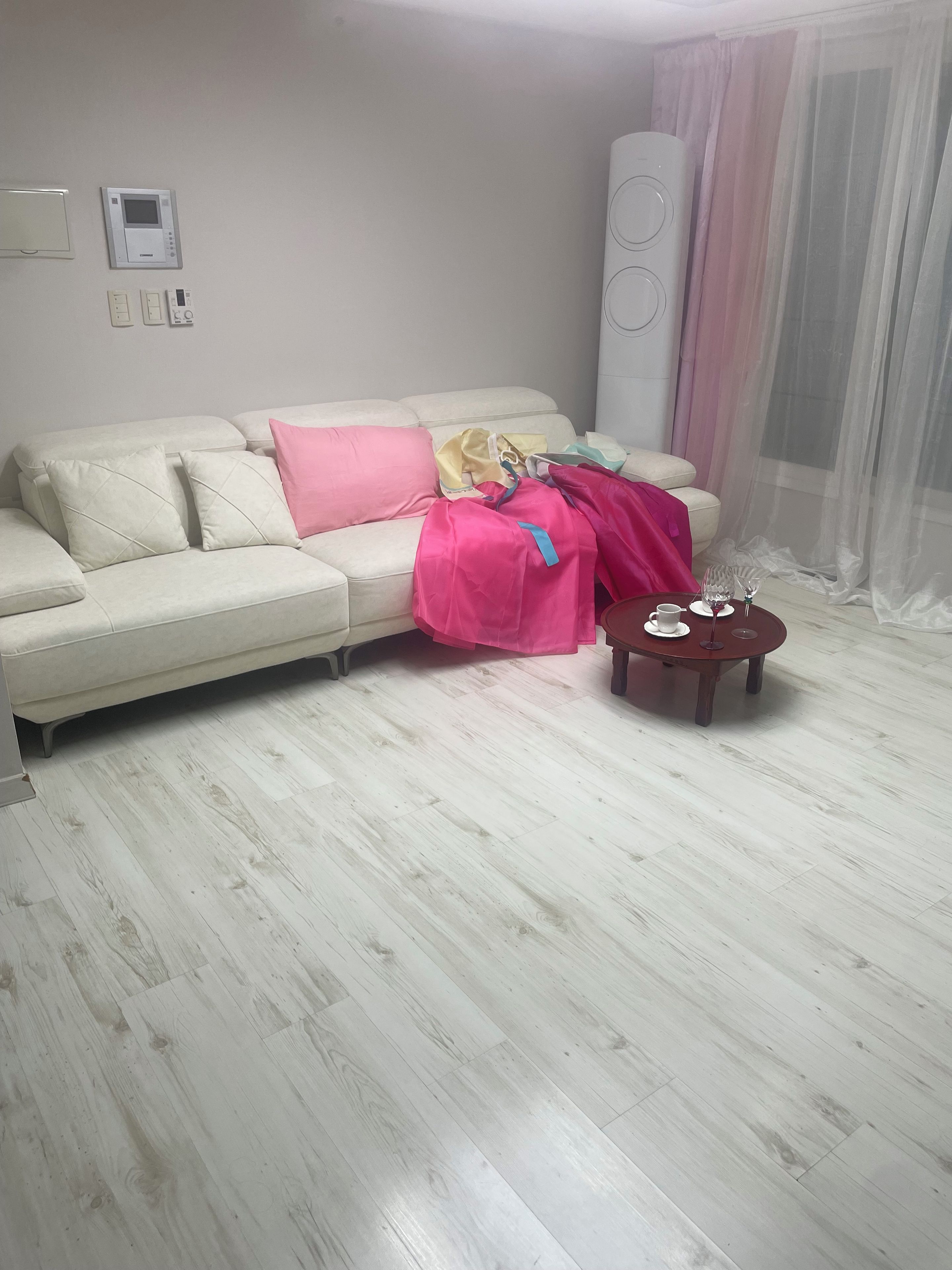 A living room with a white sofa, a pink blanket, a small wooden side table with a cup and saucer, and a large air conditioning unit on the wall.