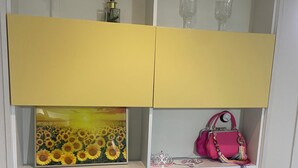 Een witte kast met verschillende vakken, waaronder een plank met een afbeelding van een zonnebloem, een roze handtas en diverse glazen voorwerpen.