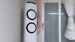 Een slaapkamer met een bed, een witte luchtreiniger, een raam met gordijn en een wandthermostaat.