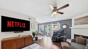 Ein Wohnzimmer mit großem Flachbildfernseher, Deckenventilator, Kamin und Essbereich mit Blick ins Freie.