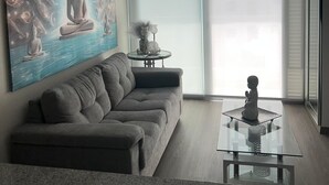 Una sala de estar moderna con un sofá, una mesa de centro de vidrio, un cuadro de Buda y un ventanal corredero que da a un balcón.
