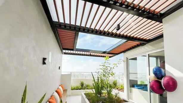 Un balcón moderno al aire libre con una pérgola de madera, una maceta con plantas y un área de descanso con cojines.