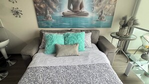 Un dormitorio con una cama grande, un cuadro colgado en la pared que muestra una figura meditando, una mesita con objetos decorativos y una silla.