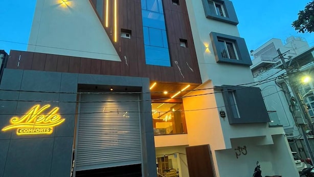 A multi-story building with a modern design, featuring a neon sign for 'Nele Comforts' and a visible entrance with steps leading up to the door. - Nele comforts (Bengaluru,)