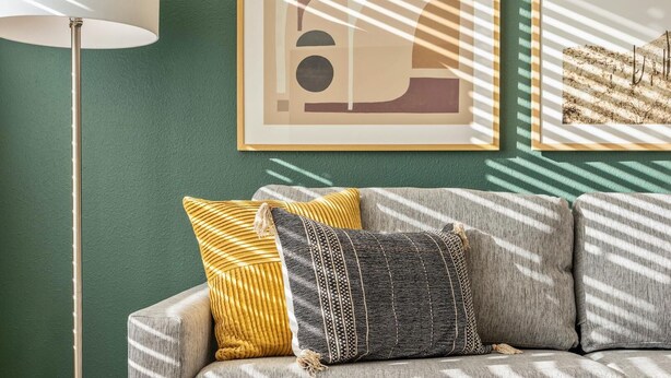 Una sala de estar con un sofá, dos almohadas, una lámpara de pie y obras de arte enmarcadas en la pared.