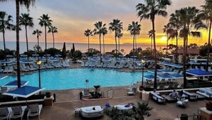 Et resort med poolområde, liggestole og parasoller, omgivet af palmer og udsigt til solnedgang.