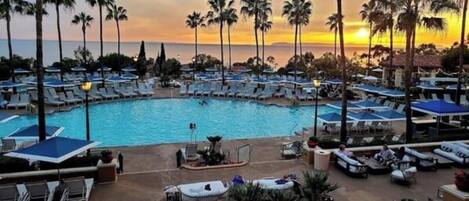 A resort pool area with lounge chairs and umbrellas, surrounded by palm trees and a sunset view.