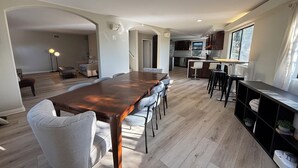 Een ruime eethoek met een lange houten tafel en meerdere stoelen, een keuken met moderne apparatuur, en een groot raam dat zorgt voor natuurlijk licht.