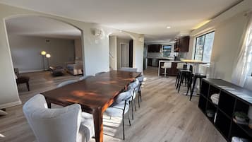 Een ruime eethoek met een lange houten tafel en meerdere stoelen, een keuken met moderne apparatuur, en een groot raam dat zorgt voor natuurlijk licht.