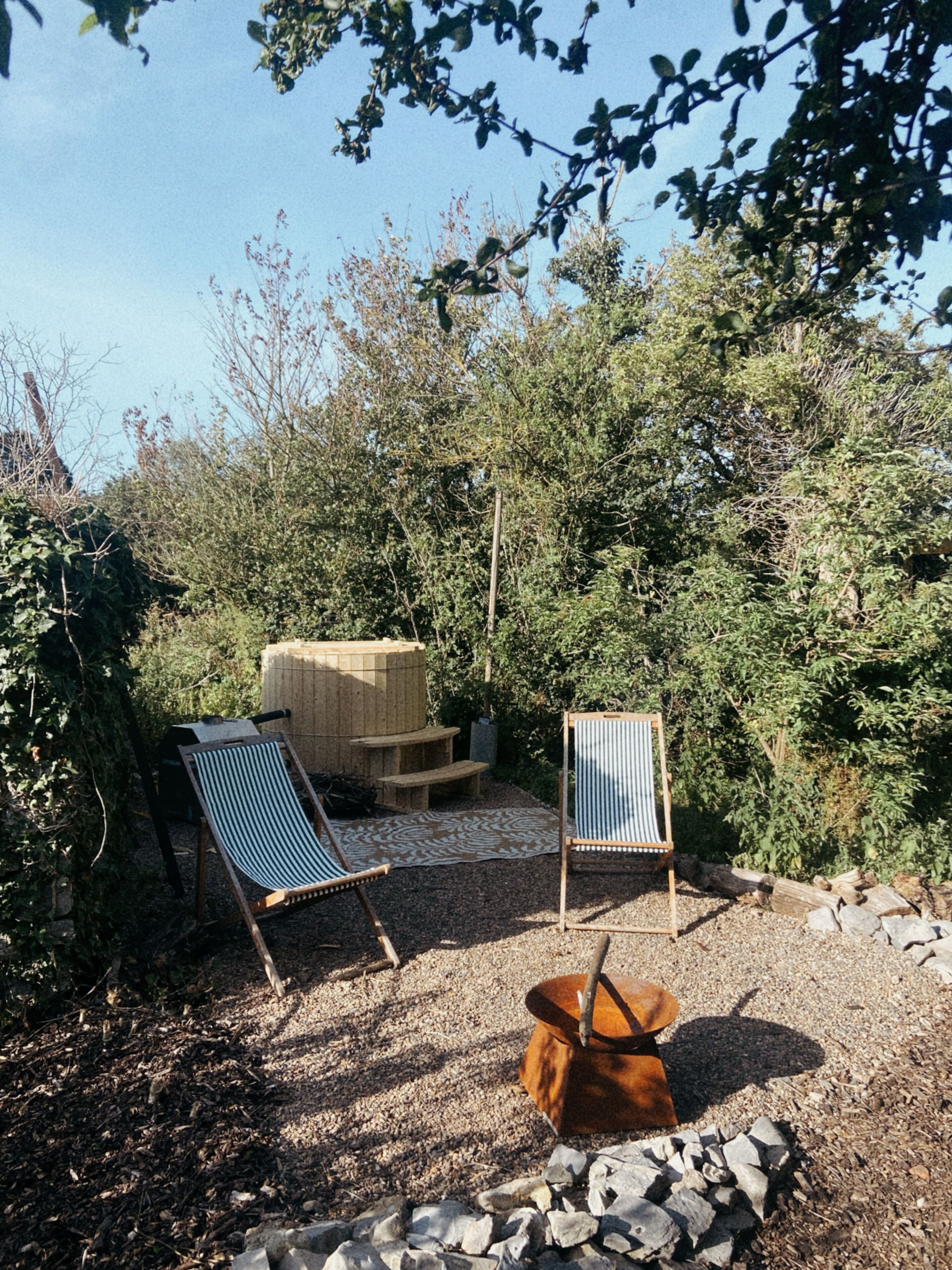 Un jardín con dos sillas de madera, un banco de madera y un pozo para fuego.