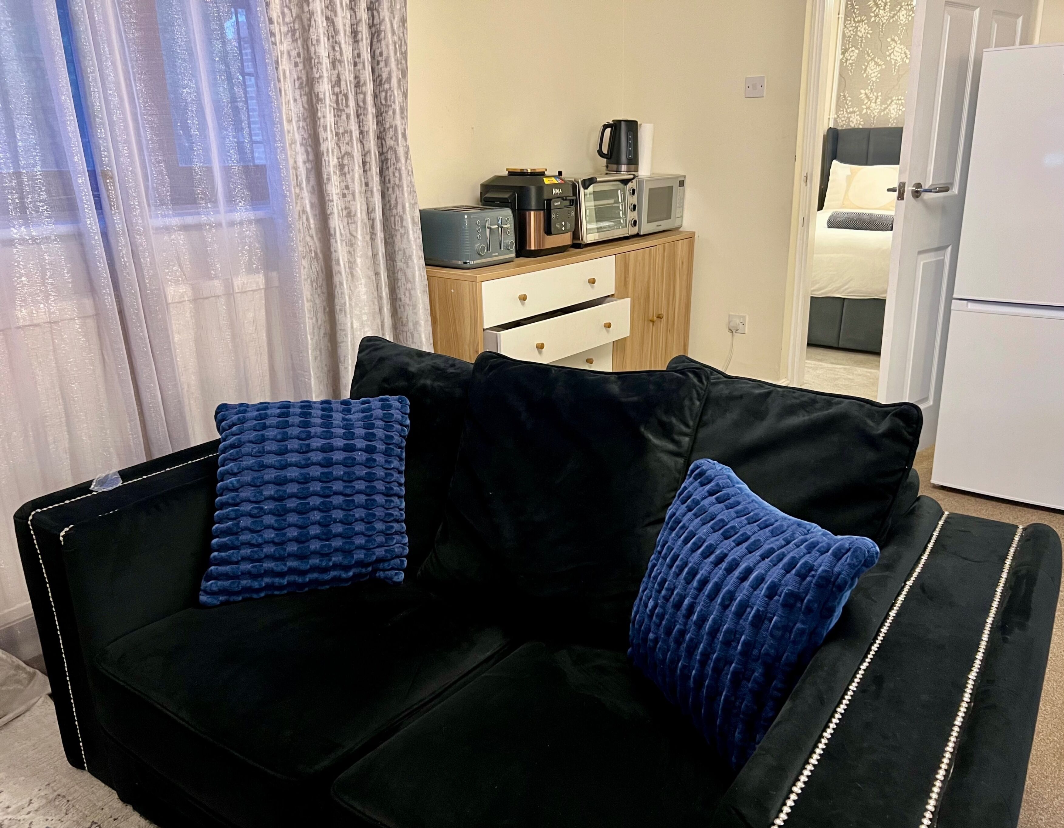 Ein schwarzes Sofa mit blauen Kissen, ein Holztischchen mit Kaffeemaschine, einer Mikrowelle und einem weißen Kühlschrank in einem Raum, der durch eine Tür mit einem weiteren Bereich verbunden ist.