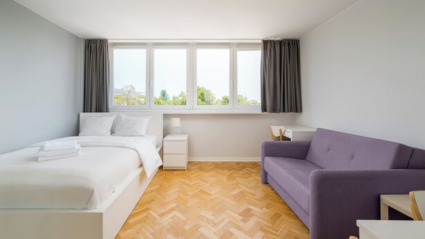 Un dormitorio moderno con cama, sofá, mesita de noche y ventana con cortinas.