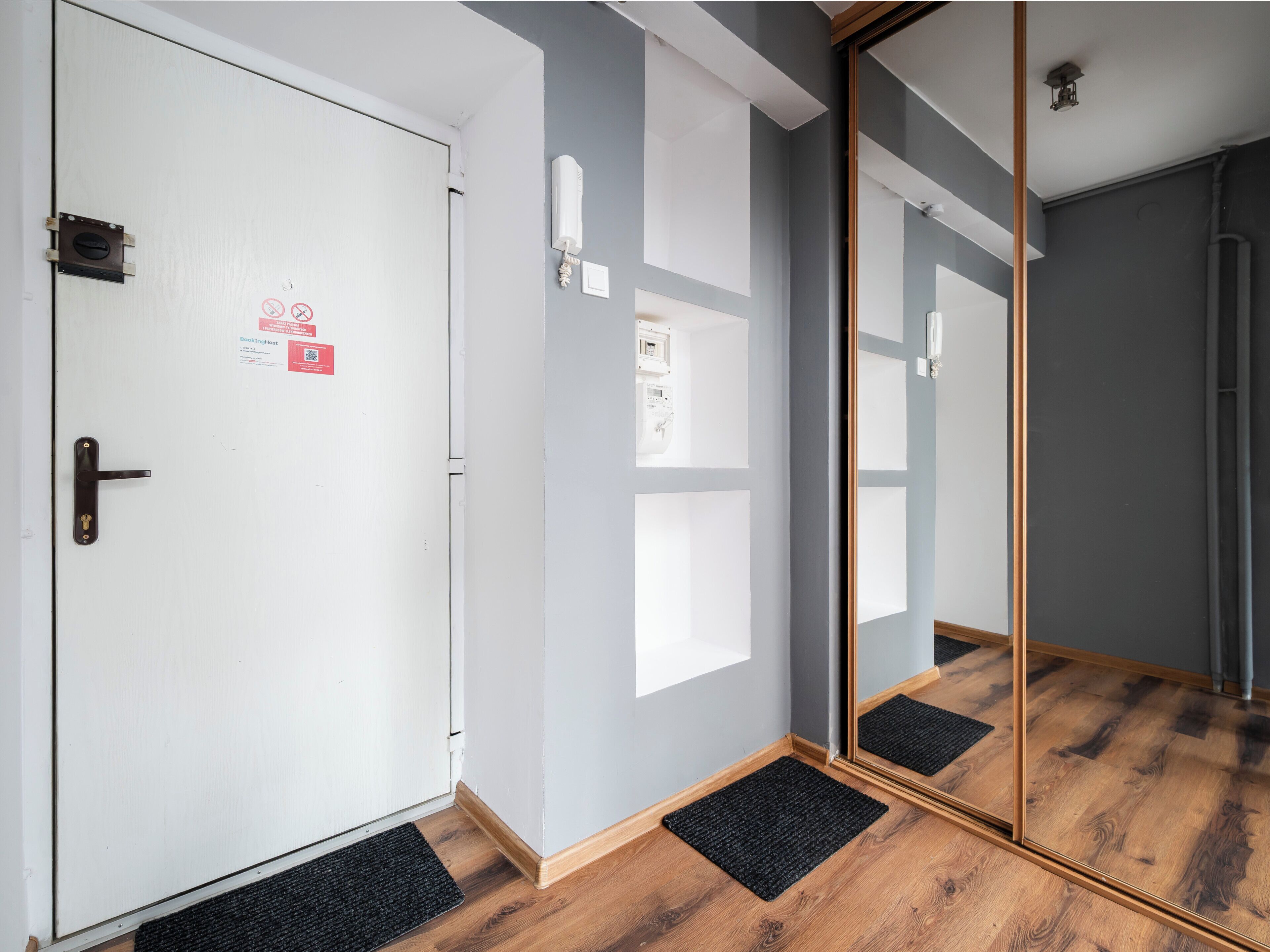 Habitación de hotel con una puerta blanca, un felpudo negro, suelo de madera y un espejo grande.