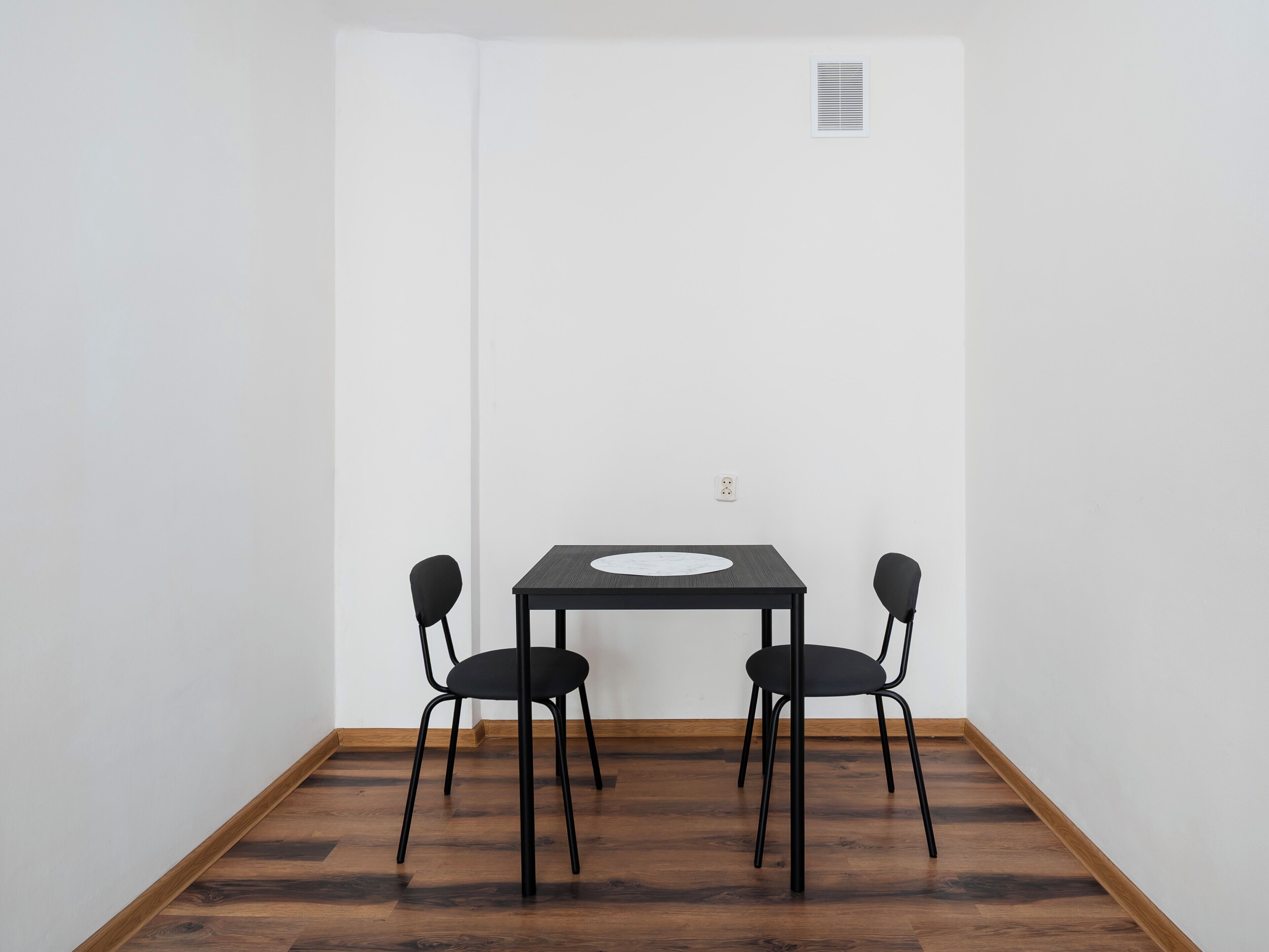 A room with a dark wooden floor, a white wall, and a black table with two chairs.