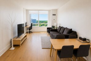 Ein modernes Wohnzimmer mit Flachbildfernseher, einer Couch, einem Esstisch mit Stühlen und einem großen Fenster mit Blick auf Gebäude und Grünflächen.