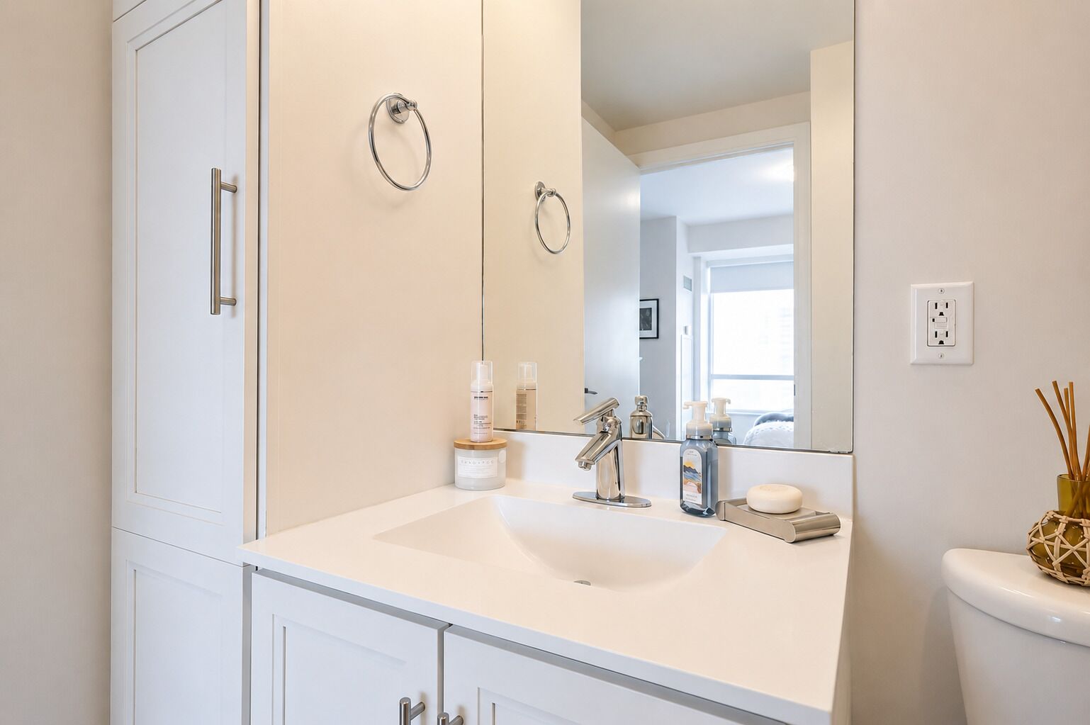 Un baño con un lavabo blanco, un espejo y un inodoro. Hay artículos de aseo en el lavabo y un florero con cañas en el inodoro.
