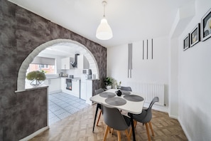 Una cocina moderna con armarios blancos, un comedor con mesa blanca y sillas grises, y un amplio arco que da a un espacio exterior.