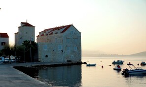 Edifici de pedra amb teulada vermella, una torre i un front marítim tranquil amb vaixells.