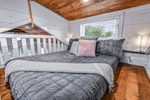 Ein Schlafzimmer mit einem großen Bett, einem Fenster mit Jalousien, einer Holzbalkendecke und einem weißen Geländer.