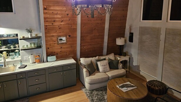 Un salon chaleureux avec un mur en bois, un lustre, un canapé, une table basse et un coin cuisine équipé de placards et d’un évier.