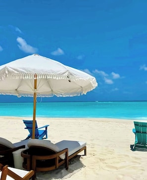 Ein Strand mit einem weißen Sonnenschirm, zwei Liegestühlen und blauem Himmel.