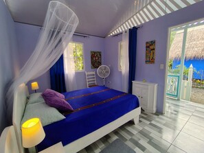 Een slaapkamer met een bed, nachtkastje, ventilator, raam en een deur naar een buitenruimte.