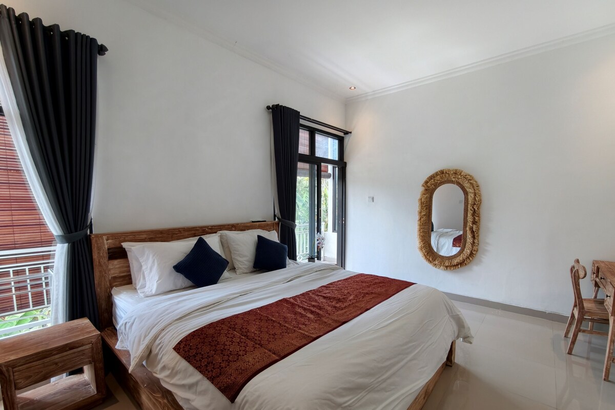 Un dormitorio con cama de madera, una manta roja, una mesita de noche de madera, un espejo y una ventana con cortinas.